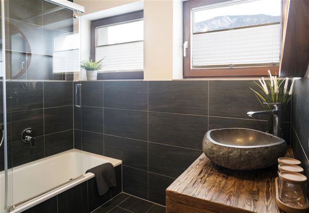 Een modern badkamer met donkere tegels en een elegante stenen wastafel. De grote douche en de ramen zorgen voor veel licht.