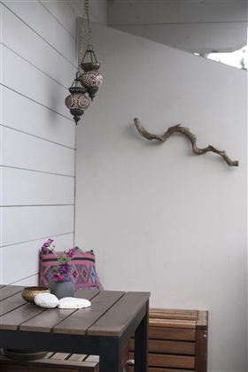 Een stijlvolle buitenruimte met een houten tafel en kleurrijke kussen. Aan de muur hangt een decoratieve lamp en een natuurlijke houten tak.