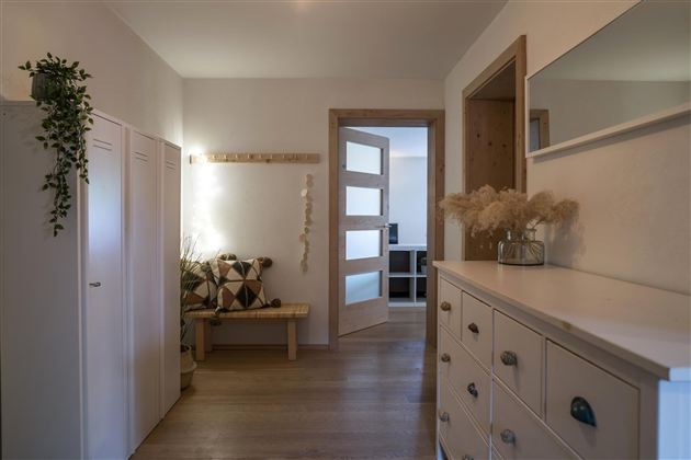 Een lichte gang met een houten vloer en een moderne inrichting. Aan de linkerkant zijn er kasten en een bank, terwijl aan de rechterkant een dressoir staat.