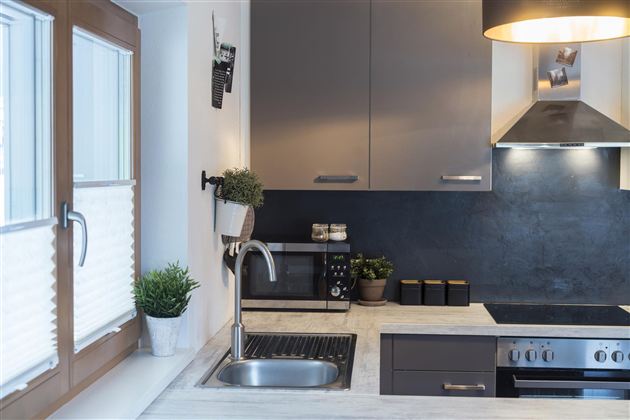 Een moderne keuken met grijze kasten en een RVS spoelbak. De ruimte is licht en smaakvol ingericht, met planten en een groot raam.