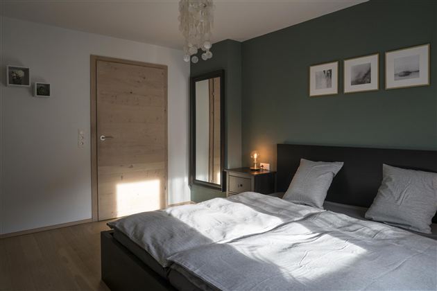 Een moderne slaapkamer met een groot bed, een spiegel en stijlvolle kunstwerken aan de muur. De kamer heeft een warme sfeer door de zonnestralen die naar binnen vallen.