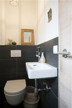 Een moderne badkamer met een stijlvolle wc en een klein wastafel. De muren zijn in een donkere tint gehouden en er zijn decoratieve elementen zoals een spiegel en een fles.