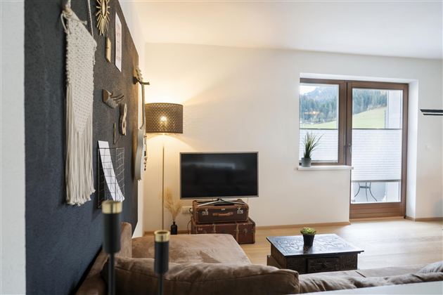 Een gezellige woonkamer met een donkere muur, een televisie en een raam met uitzicht op groen. De inrichting is modern en uitnodigend, met een bank en een tafel.
