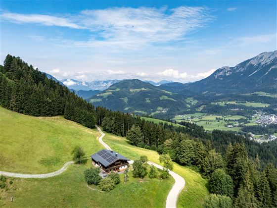 Eine malerische Landschaft mit grünen Wiesen und bewaldeten Bergen. Im Vordergrund steht ein gemütliches Haus am Fuß eines Hügels.