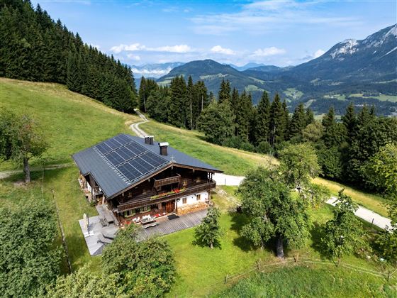Ein charmantes Haus in den Bergen umgeben von grüner Natur und hohen Bäumen. Der Himmel ist klar und es ist eine ruhige, einladende Landschaft.