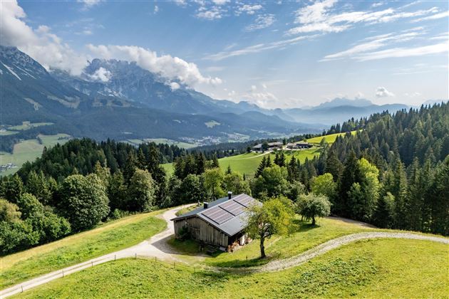 Eine malerische Landschaft mit einem Holzhäuschen auf einer Wiese. Im Hintergrund sind beeindruckende Berge und ein klarer Himmel zu sehen.