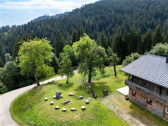 Eine malerische Landschaft mit einem Holzhaus und grünen Wiesen. Um das Haus herum stehen Bäume, und auf der Wiese gibt es einen Steinkreis.