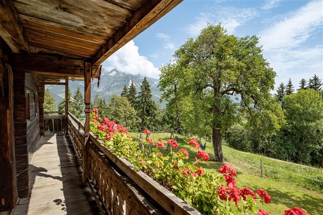 Ein Balkon mit Holzgeländer, geschmückt mit roten Blumen. Im Hintergrund sind grüne Wiesen und Berge unter einem blauen Himmel zu sehen.