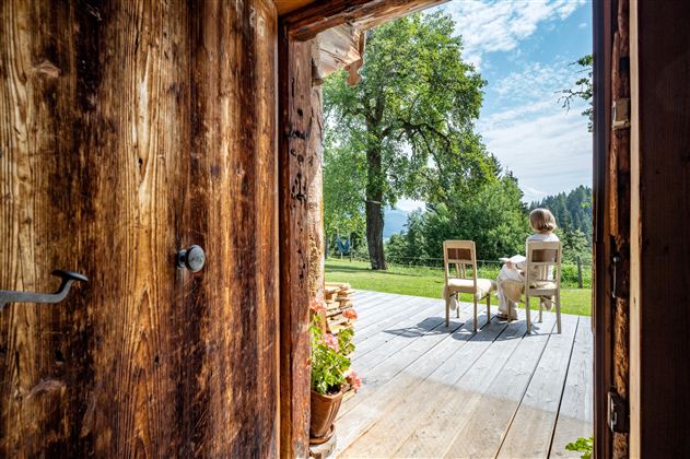 Eine offene Holztür zeigt eine Terrasse mit zwei Stühlen und einer Person, die den Ausblick genießt. Im Hintergrund sind grüne Bäume und ein klarer Himmel zu sehen.
