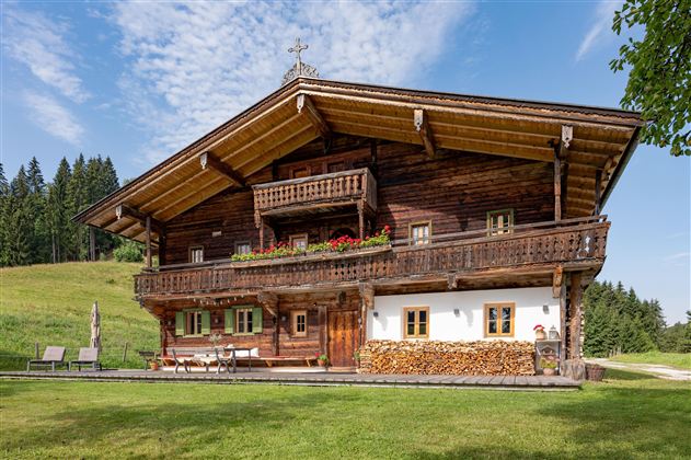 Ein traditionelles Holzhaus im alpinen Stil mit einem Balkon und bunten Blumen. Umgeben von grünen Wiesen und Bäumen unter einem klaren blauen Himmel.