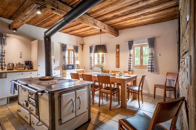 Eine gemütliche Küche mit rustikalem Design und Holzbalkendecke. Ein großer Esstisch steht im Mittelpunkt, umgeben von Stühlen und Fenster mit Vorhängen.