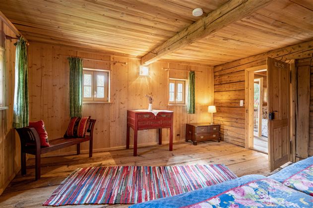 Ein gemütliches Holzzimmer mit buntem Teppich und natürlichem Licht. Es gibt einen kleinen Tisch, Stühle und Zugang zur Außentür.