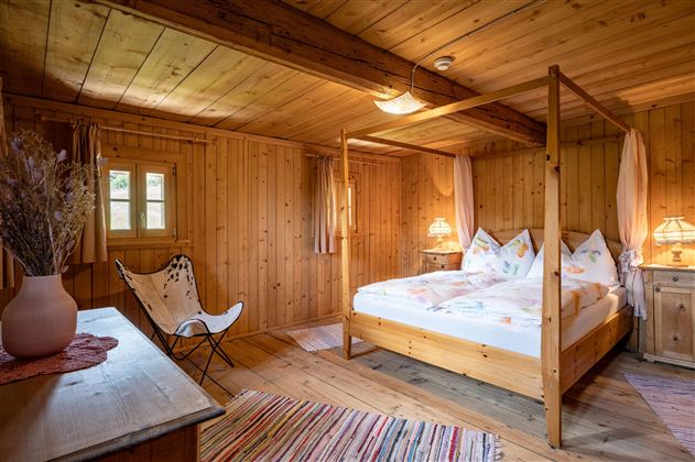Ein gemütliches Schlafzimmer mit Holzwänden und einem Himmelbett. Der Raum ist warm und einladend, mit sanftem Licht und einfachen Möbeln.