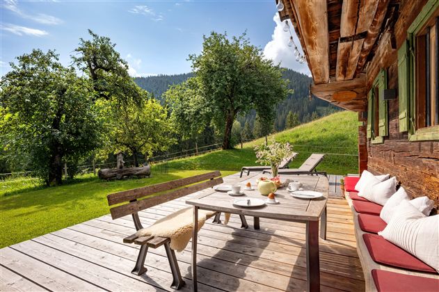 Eine gemütliche Terrasse mit einem Holztisch und Bänken, umgeben von grünen Bäumen und einer malerischen Berglandschaft. Die Sonne scheint und lädt zum Entspannen im Freien ein.