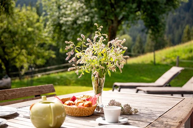 Ein Tisch im Freien, gedeckt mit einer Teekanne, einem Becher und einer Obstschale. Im Hintergrund sind grüne Wiesen und Bäume zu sehen, was eine entspannte Atmosphäre schafft.
