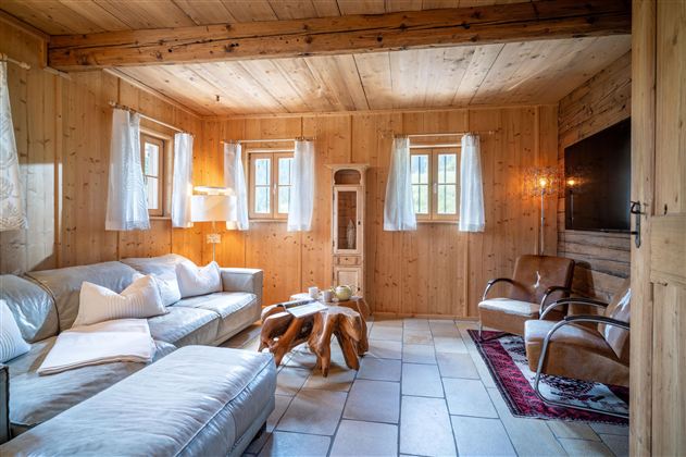 Ein gemütliches Wohnzimmer mit Holzverkleidung und große Fenster. Der Raum ist mit einem hellen Sofa und zwei Sesseln eingerichtet.