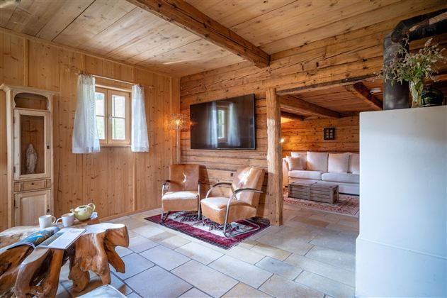 Ein gemütlicher Wohnraum mit Holzwänden und viel natürlichem Licht. Zwei Ledersessel stehen vor einem Fernseher und einem Sichtbereich mit einer gemütlichen Couch.
