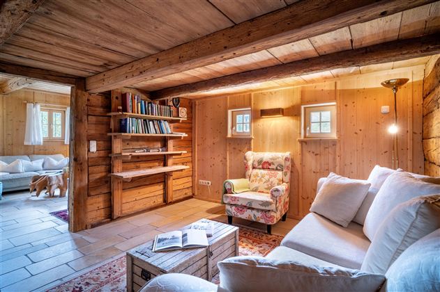 Ein gemütliches, rustikales Wohnzimmer mit Holzwänden und einem bequemen Sessel. Regale mit Büchern und sanftem Licht schaffen eine einladende Atmosphäre.
