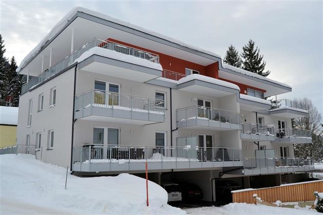 A modern apartment building in winter, surrounded by snow. The balconies are clearly visible and there are cars in the garage below.