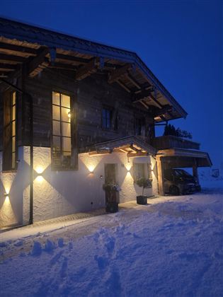 A cozy wooden house in the snow, illuminated by warm lights. The evening sky is dark blue and fresh snow lies on the ground.