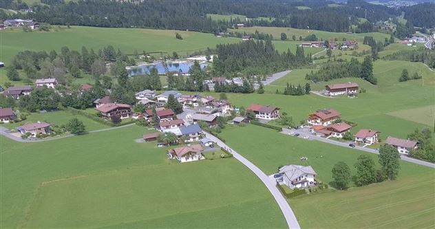 A picturesque landscape with a small village, surrounded by green meadows and forests. In the background, several buildings and a swimming pond can be seen.