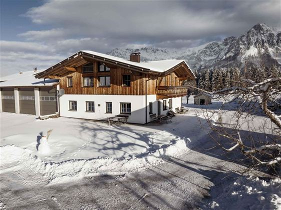 Ein gemütliches Ferienhaus in den Bergen, umgeben von frisch gefallenem Schnee. Im Hintergrund sind majestätische Berge und ein klarer Himmel zu sehen.