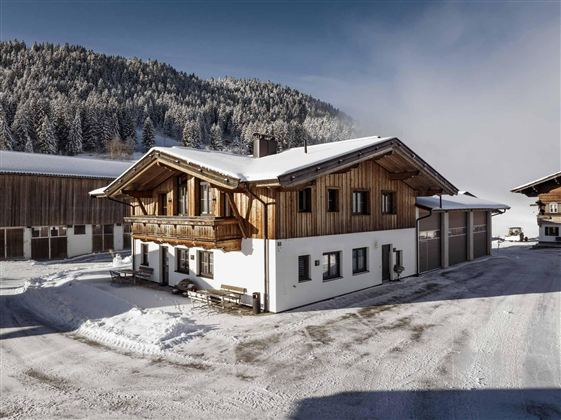 Ein traditionelles Holzhaus in einer schneebedeckten Landschaft. Im Hintergrund sind bewaldete Berge zu sehen.