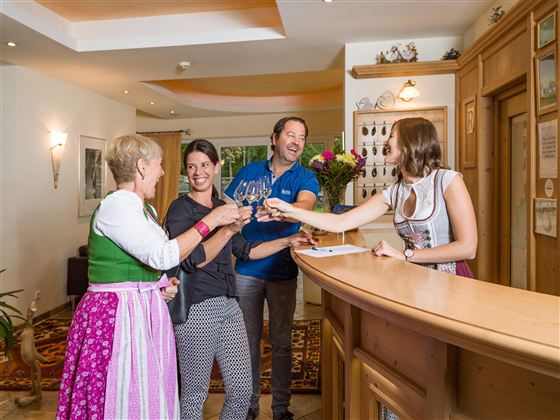 Eine fröhliche Gruppe von Menschen stößt an der Rezeption eines Hotels an. Die Gäste tragen traditionelle Kleidung und genießen einen herzlichen Empfang.