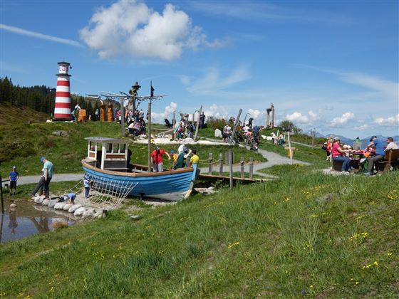 Ein malerischer Aussichtspunkt mit einem Boot und einem Leuchtturm im Hintergrund. Viele Menschen genießen die Natur und sitzen auf der Wiese.