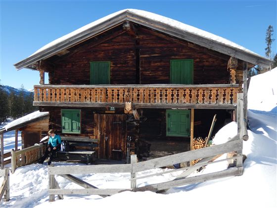 Eine traditionelle Berghütte aus Holz mit grünen Fensterläden steht im Schnee. Der Himmel ist klar und blau, und die Umgebung ist winterlich.
