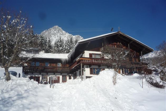 Ein traditionelles Chalet im Schnee, umgeben von Bäumen. Im Hintergrund ist ein schneebedeckter Berg und ein strahlend blauer Himmel zu sehen.