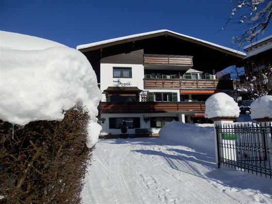 Ein gemütliches Holzhaus im Schnee mit Balkonen und einem strahlend blauen Himmel. Der Weg vor dem Haus führt durch eine verschneite Landschaft.