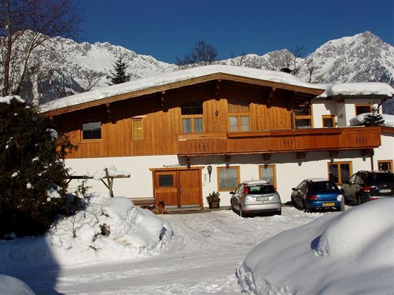 A wooden house with a balcony, surrounded by snow and mountains. In the foreground, several cars are parked in a snowy parking lot.