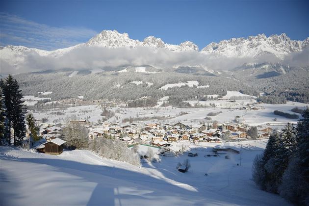 Eine winterliche Landschaft mit schneebedeckten Bergen im Hintergrund und einem kleinen Dorf im Tal. Die Bäume und Felder sind ebenfalls mit Schnee bedeckt, was eine ruhige und friedliche Atmosphäre schafft.
