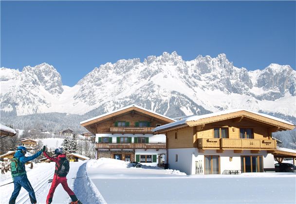 Two skiers are enjoying the snow in front of chalets in a winter mountain landscape. In the background, majestic mountains rise beneath a radiant blue sky.