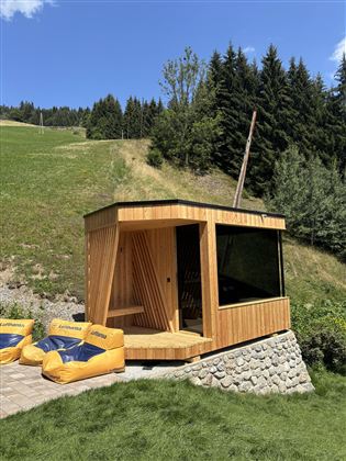 Eine moderne Holzsauna steht in der Natur, umgeben von Bäumen und Wiese. Vor der Sauna liegen zwei gelbe Liegestühle.