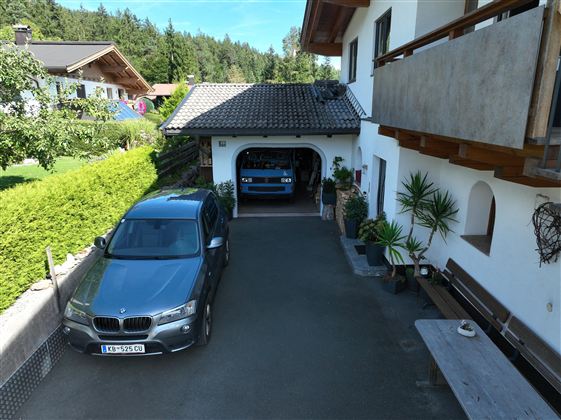 Eine Auffahrt mit einem grauen Auto vor einer Garage, in der ein weiteres blaues Auto steht. Im Hintergrund sind grüne Bäume und ein Wohnhaus zu sehen. Das zeigt die Parksituation der Gäste ein Garagen und Aussen Stellplatz.