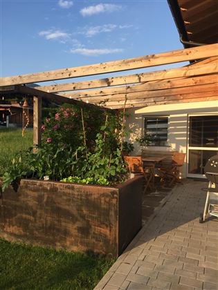 Eine Terrasse mit einer Holzpergola und einem Gartenbeet. Im Hintergrund ist ein schattiges Plätzchen mit Gartenmöbeln und einem Grill.
