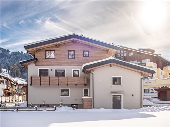 Ein modernes Haus im Schnee mit einer Holzveranda. Im Hintergrund sind Berge und weitere Gebäude zu sehen.