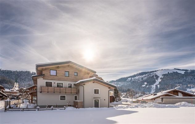 Ein modernes Haus in einer verschneiten Landschaft. Die Sonne scheint über den Bergen und dem schneebedeckten Boden.