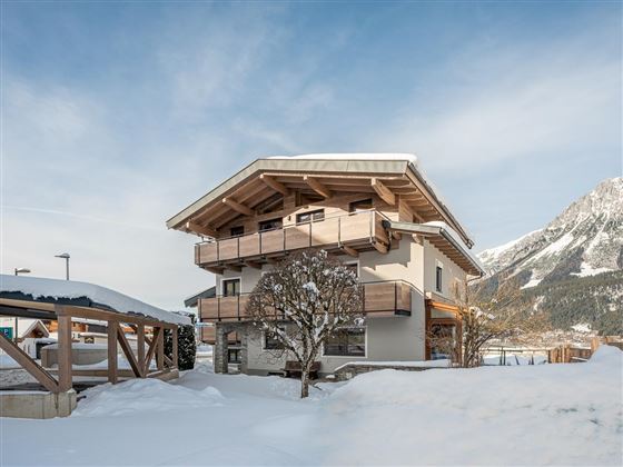 Ein modernes Haus im Schnee mit Holzverkleidung und Balkonen. Im Hintergrund sind Berge unter einem klaren Himmel zu sehen.