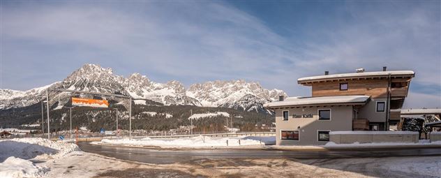 Eine malerische Winterlandschaft mit schneebedeckten Bergen im Hintergrund. Im Vordergrund steht ein modernes Gebäude an einer gut ausgebauten Straße.