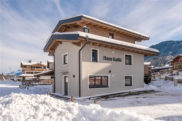 Ein modernes Haus im Schnee mit dem Namen "Haus Kathi" an der Fassade. Im Hintergrund sind Berge und weitere Gebäude zu sehen.