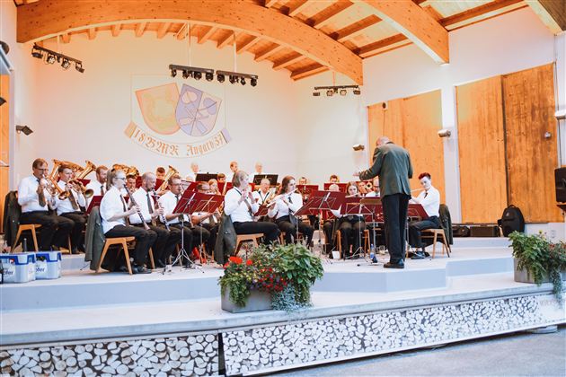 A brass band is playing on a stage in a cozy indoor setting. The conductor is leading the musicians, while some plants are decoratively placed.
