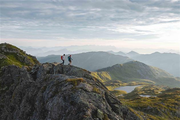 Twee wandelaars staan op een rotsachtige top en kijken uit over het pittoreske berglandschap. Op de achtergrond rijzen zachte heuvels en majestueuze bergen onder een bewolkte lucht.