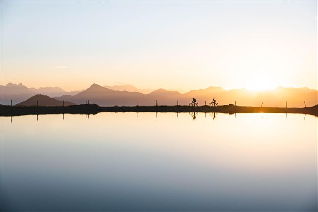 Twee fietsers rijden langs een rustige waterweg bij zonsondergang. De bergen op de achtergrond creëren een indrukwekkend uitzicht.