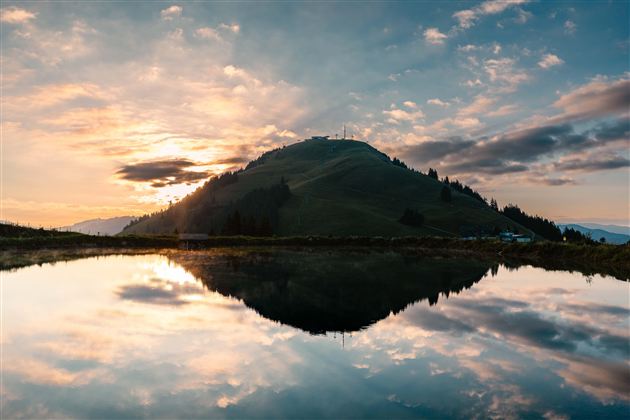 Een pittoresk landschap met een heuvel op de achtergrond en een rustige meer op de voorgrond. De lucht is gevuld met wolken en een mooie zonsondergang.