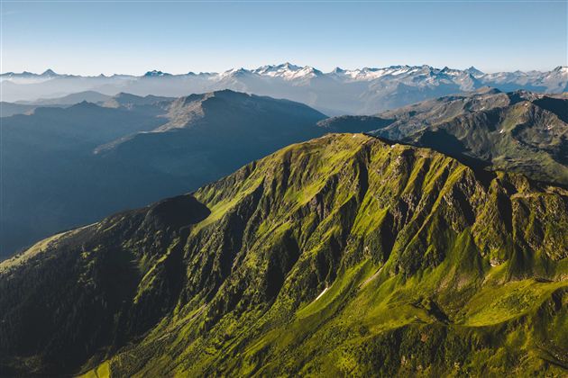 Een indrukwekkend berglandschap met groene heuvels en besneeuwde toppen op de achtergrond. De lucht is helder en er is een vredige sfeer te voelen.