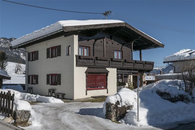 A cozy house in the snow with an upper balcony. The landscape is wintry and the mountains are visible in the background.