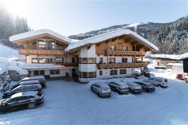A traditional building in the Alps, surrounded by snow-covered fir trees. In the foreground, several cars are on the snow-covered parking lot.